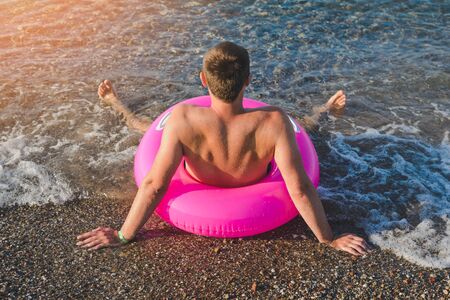 Happy man in pink with a big inflatable ring on the sea beach in summer sunny day. Back view.の写真素材