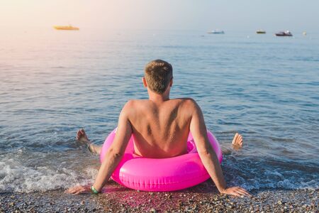 Happy man with big pink inflatable ring on the sea beach in summer sunny day. Back view.の写真素材