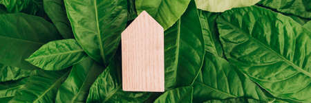 Banner with miniature wooden house on green leaves. Eco-friendly and energy efficient house concept.