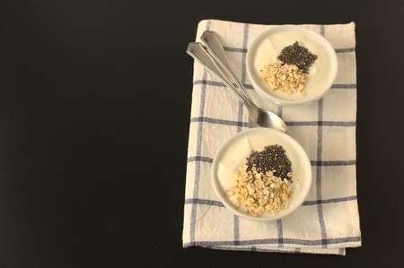 Two bowls with greek yogurt, chi a seeds and oatmeal, next to two teaspoon on white and blue plaid clothの写真素材