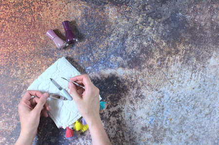 Woman doing her nails with a manicure set consisting of cuticle pusher, nail clipper and file on a white towel with colored tassels, next to it some nail polish, on a rusty-looking background.の写真素材
