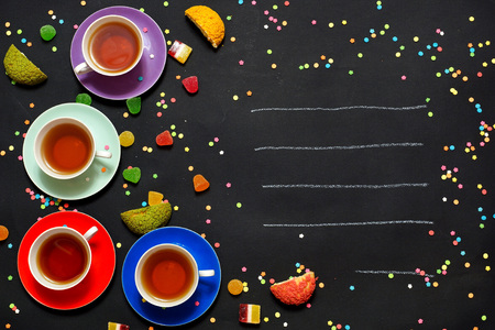 Four colorful cups of tea, marmalade and biscuits on a black background. Free space for text. A festive greeting card with cups and sweets. View from aboveの写真素材