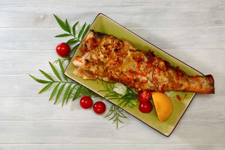 Baked fish with vegetables in a green plate on a white table decorated with herbs and tomatoes