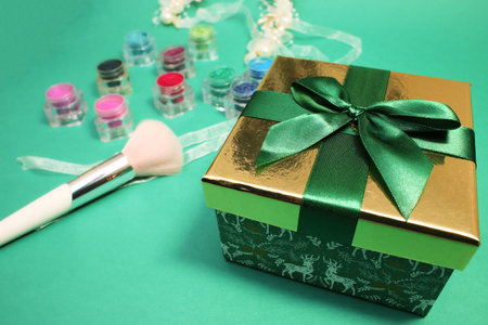 Green gift box with gold cover and green ribbon bow and professional makeup brush and rainbow eye shadow on green background.