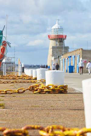 Stone pier with big white bollard and yellow metal chain for ships and white lighthouse.の写真素材