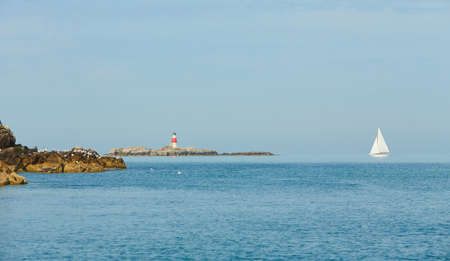 Old Muglins Lighthouse on the isolated island the backdrop of the blue cloudy sky, calm water and...