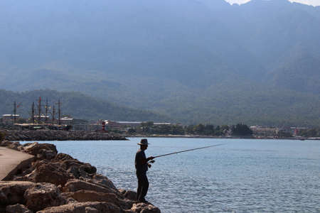 A fisherman with a fishing rod on the seashore against the background of mountains.の写真素材