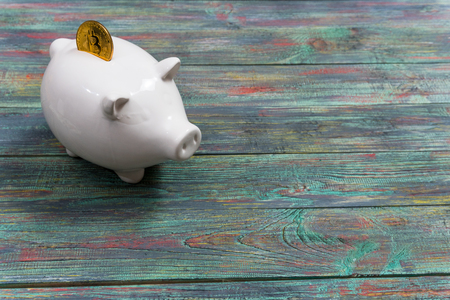 White piggy bank with one gold bitcoin coin new virtual money on a colored wooden backgroundの写真素材