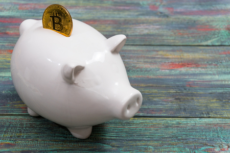 White piggy bank with one gold bitcoin coin new virtual money on a colored wooden backgroundの写真素材