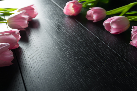 Border from bright pink tulips flowers on black wooden background. Selective focus. Place for text. Flat lay. With an artificial Sun Reflexの写真素材