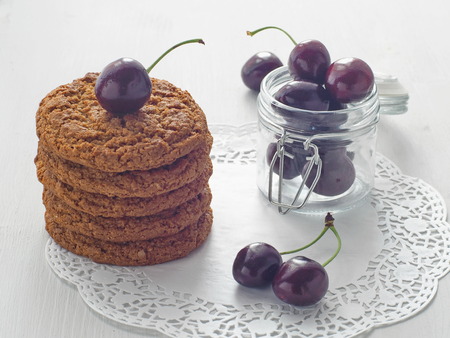 Pile of homemade oatmeal cookies decorated with ripe cherry with a jar full of fresh cherries on white wooden table. Selective focus.の写真素材