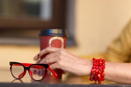 Female hand on a table in a cafe in a red bracelet, with a paper cup for coffee and glasses on the tableの写真素材
