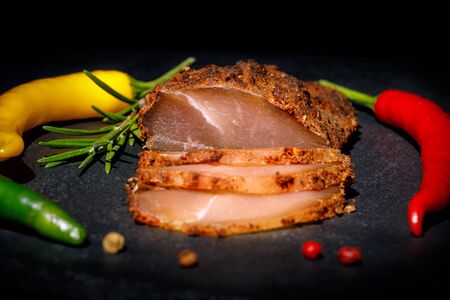 Chicken breast basturma on a black background with colored chili peppers and a sprig of rosemaryの写真素材