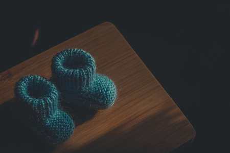 A pair of knitted blue handmade baby booties on a wooden board. Close-up, copy space, horizontal composition, dark background, deep shadowsの写真素材