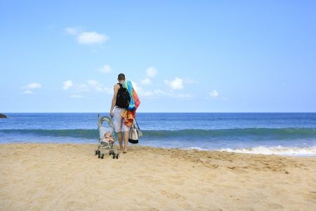 Hands full during vacation with baby  Young father is dragging stroller with little baby on the sandy beach near ocean の写真素材