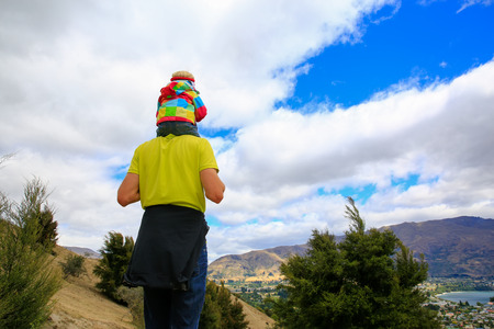 Young man with a little girl on his shoulders staying on the top of the mountain and appreciating beautiful lake Wanaka landscape, New Zealandの写真素材