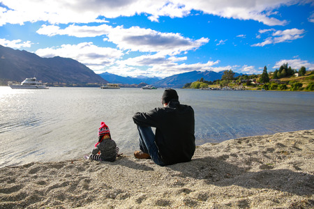 Father with little daughter sitting on sandy beach, appreciating Lake Wanaka landscape in Otago region, South Island, New Zealandの写真素材