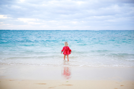 Little girl in red coat staying in the water on the beach with blue water background. Oahu , New Zealandの写真素材