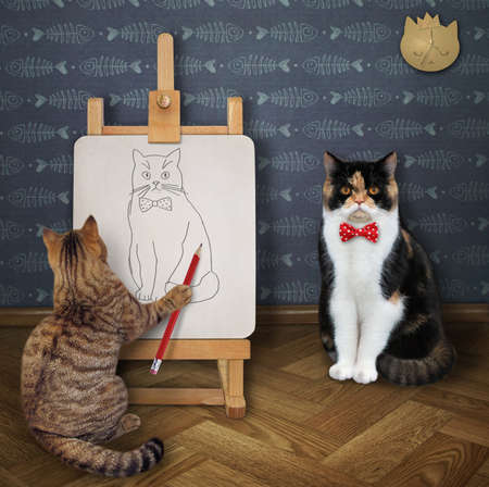 A beige cat artist with a paintbrush paints his friend in a bow tie on a canvas on an easel in his art studio.の写真素材