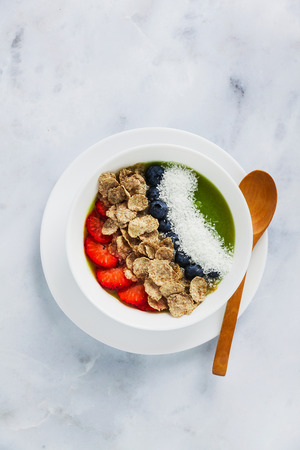 a bowl of smoothies, with cereals for a healthy breakfast and berries, chia seeds and coconut chips freshly sliced fruit, Wheat bran and Cornflake. Served with a wooden spoon on a white marble tableの写真素材