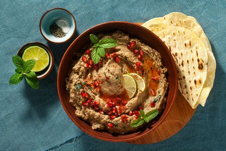 homemade spread of baked eggplant baba ganoush in a bowl with pomegranate seeds, lime, olive oil and lime slices on a blue linen tablecloth with flat breadの写真素材