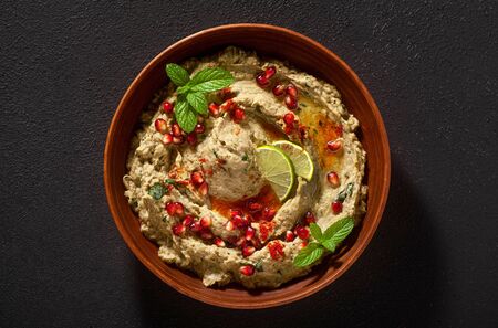 homemade spread of baked eggplant baba ganoush in a bowl with pomegranate seeds, lime, olive oil and lime slices on a black backgroundの写真素材