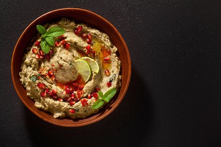 homemade spread of baked eggplant baba ganoush in a bowl with pomegranate seeds, lime, olive oil and lime slices on a black backgroundの写真素材