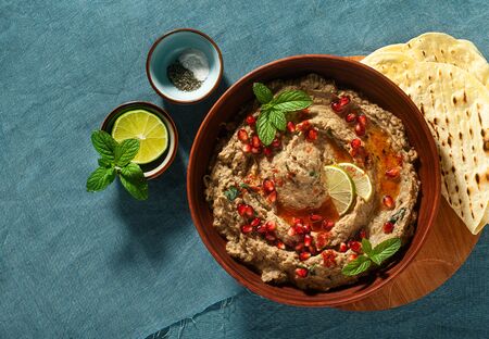 homemade spread of baked eggplant baba ganoush in a bowl with pomegranate seeds, lime, olive oil and lime slices on a blue linen tablecloth with flat breadの写真素材