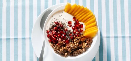 banner of breakfast bowl of granola, yogurt, fresh sweet mango, chia seeds, pomegranate seeds and maple syrup on a blue striped napkinの写真素材