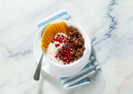 breakfast bowl of granola, yogurt, fresh sweet mango, chia seeds, pomegranate seeds and maple syrup on a blue striped napkinの写真素材