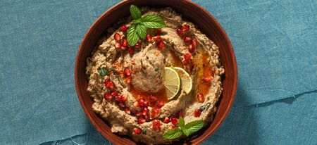 banner of homemade spread of baked eggplant baba ganoush in a bowl with pomegranate seeds, lime, olive oil and lime slices on a blue linen tableclothの写真素材