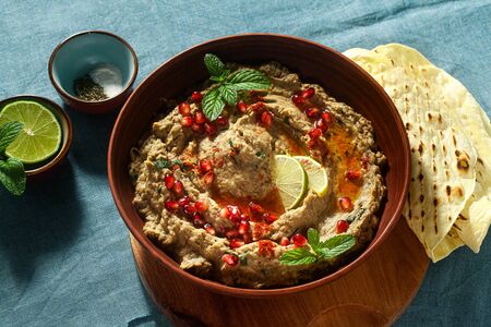 homemade spread of baked eggplant baba ganoush in a bowl with pomegranate seeds, lime, olive oil and lime slices on a blue linen tablecloth with flat breadの写真素材