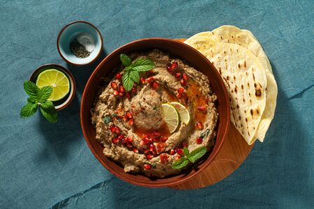 homemade spread of baked eggplant baba ganoush in a bowl with pomegranate seeds, lime, olive oil and lime slices on a blue linen tablecloth with flat breadの写真素材