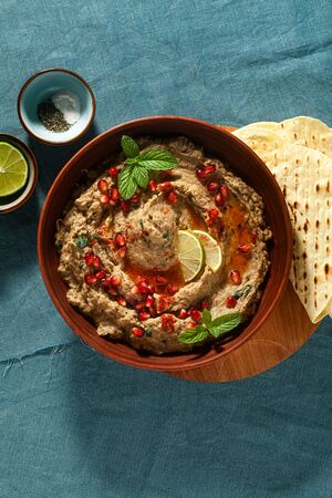 homemade spread of baked eggplant baba ganoush in a bowl with pomegranate seeds, lime, olive oil and lime slices on a blue linen tablecloth with flat breadの写真素材