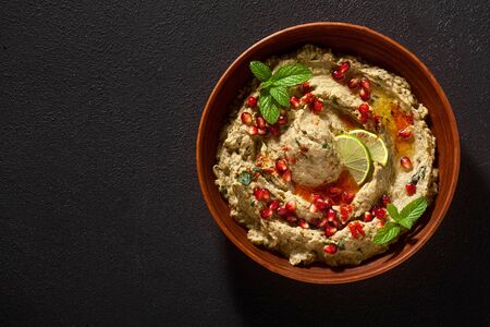 homemade spread of baked eggplant baba ganoush in a bowl with pomegranate seeds, lime, olive oil and lime slices on a black backgroundの写真素材