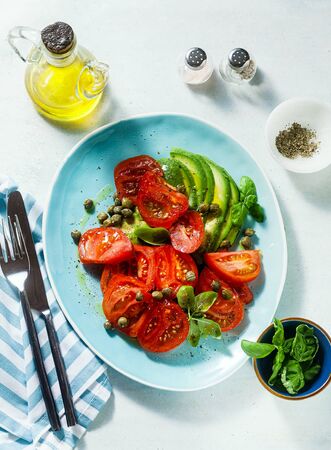 summer salad with ripe tomatoes and avocado with capers in a blue plate on the tableの写真素材