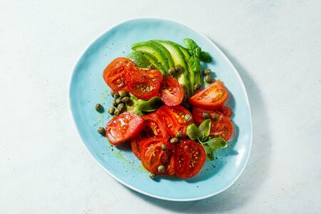 summer salad with ripe tomatoes and avocado with capers in a blue plate on the tableの写真素材
