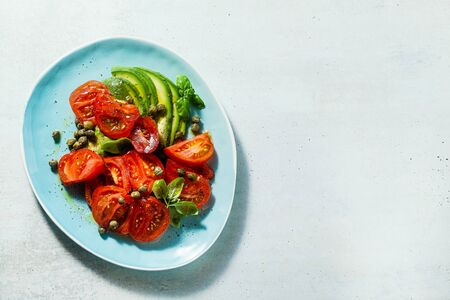 summer salad with ripe tomatoes and avocado with capers in a blue plate on the tableの写真素材