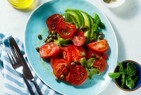 summer salad with ripe tomatoes and avocado with capers in a blue plate on the tableの写真素材