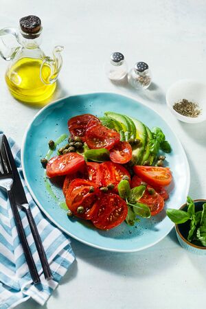 summer salad with ripe tomatoes and avocado with capers in a blue plate on the tableの写真素材