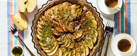 banner of italian apple pie crostata with pistachio crumbs on the table with a white tablecloth and cups of coffee espressoの写真素材