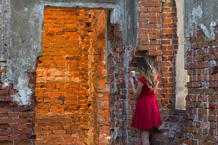 Pretty girl in red dress in the middle of destroyed brick walls with smartphone in her hands