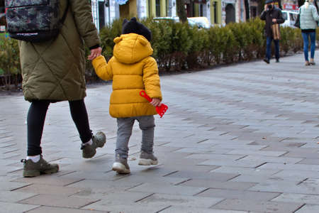 Ukraine Zhytomyr March 29, 2021. Kid in bright yellow jacket walking with mother by the hand on sidewalk on blurred background of city street. Back viewのeditorial素材