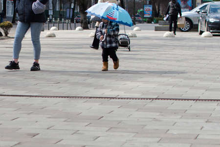 Ukraine Zhytomyr March 29, 2021. Kid under umbrella walking with his mother on sidewalk in sunny day on blurred background of city streetのeditorial素材