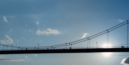 Silhouette of pedestrian bridge with people on background of blue sky with clouds and bright sun