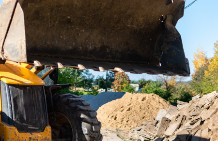Excavator bucket close-up on blurred background of construction site with pile of sand and old stone blocksの写真素材