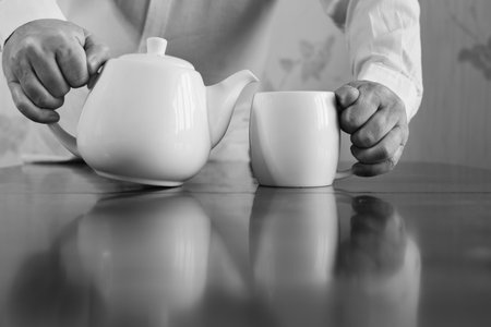 Close-up hands of middle-aged man with white ceramic teapot and white ceramic mug on wooden table on neutral blurred background. Black and white imageの写真素材