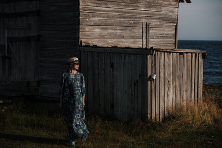 A woman is standing in front of a small, old building. The building is made of wood and has a slanted roof. The woman is wearing a dress and a hat. The sky is clearの写真素材