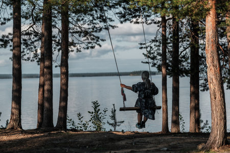 A woman is swinging on a tree swing in a forest near a lake. The scene is peaceful and serene, with the sound of the water and the rustling of the trees in the backgroundの写真素材