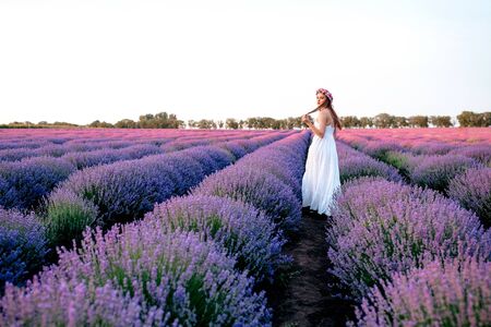 Beautiful woman in a white dress with a pink wreath walks on a lavender fieldの写真素材